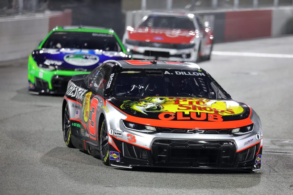 A Chevrolet NASCAR vehicle featuring a bold design with the number 3 and sponsorship from Bass Pro Shops, racing on a track at night, with two other cars in the background.