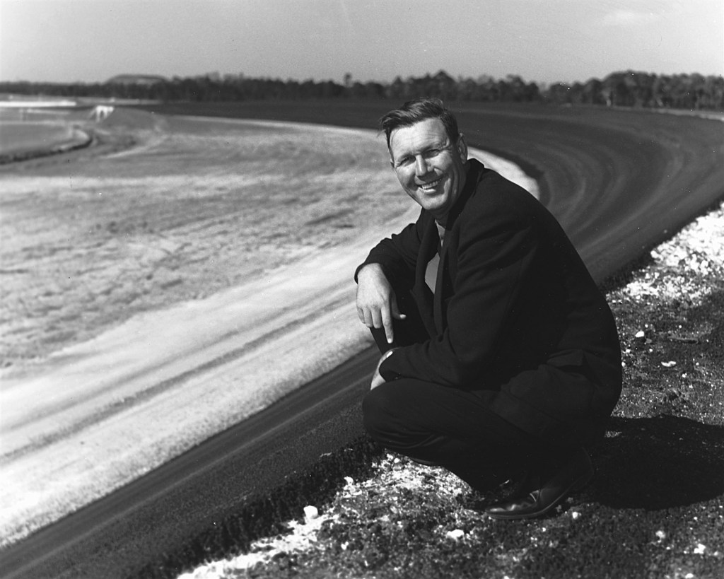 Black and white photo of a man in a suit smiling while crouched near a racetrack, with a curved dirt path and trees in the background.