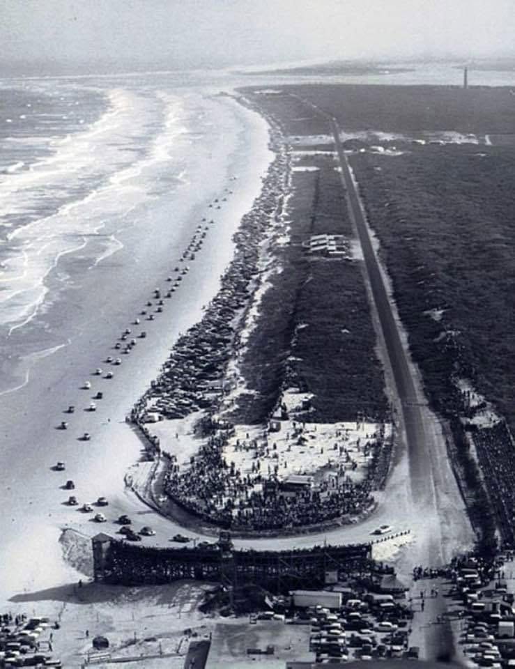 Aerial view of a crowded beach with cars parked along the shoreline, featuring a winding road and a structure near the water, likely a pier or boardwalk.