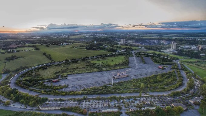 Aerial view of an abandoned industrial area featuring an empty racetrack surrounded by greenery and overgrown vegetation, with distant buildings and a sunset sky in the background.
