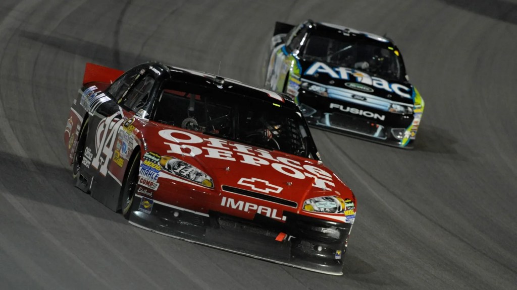 Two race cars competing on a track at night, with one car in the foreground featuring a red paint job and the 'Office Depot' logo, while the other car, slightly behind, is blue with 'Aflac' branding.